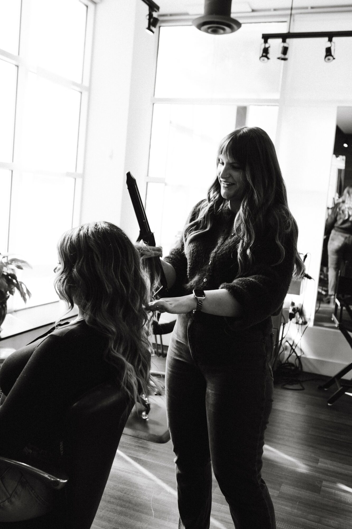 Two women in a bright room, one standing and styling the other's hair, with large windows and modern decor.