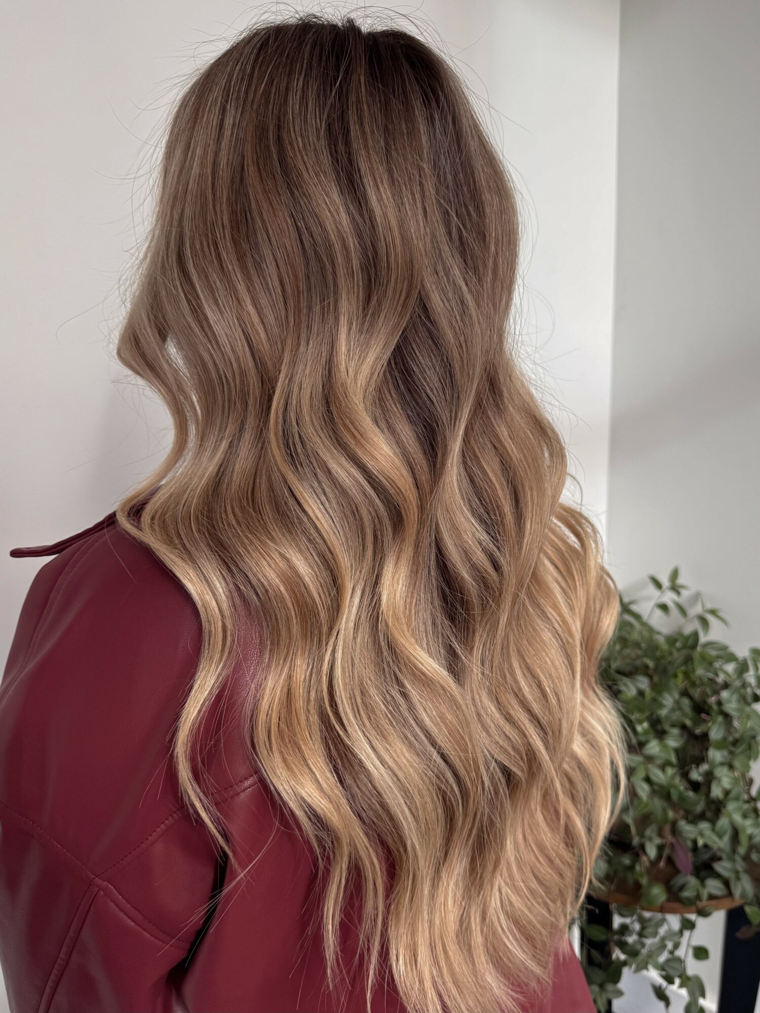 Back view of a woman with long, wavy, light brown hair with blonde highlights, wearing a maroon top, standing indoors.