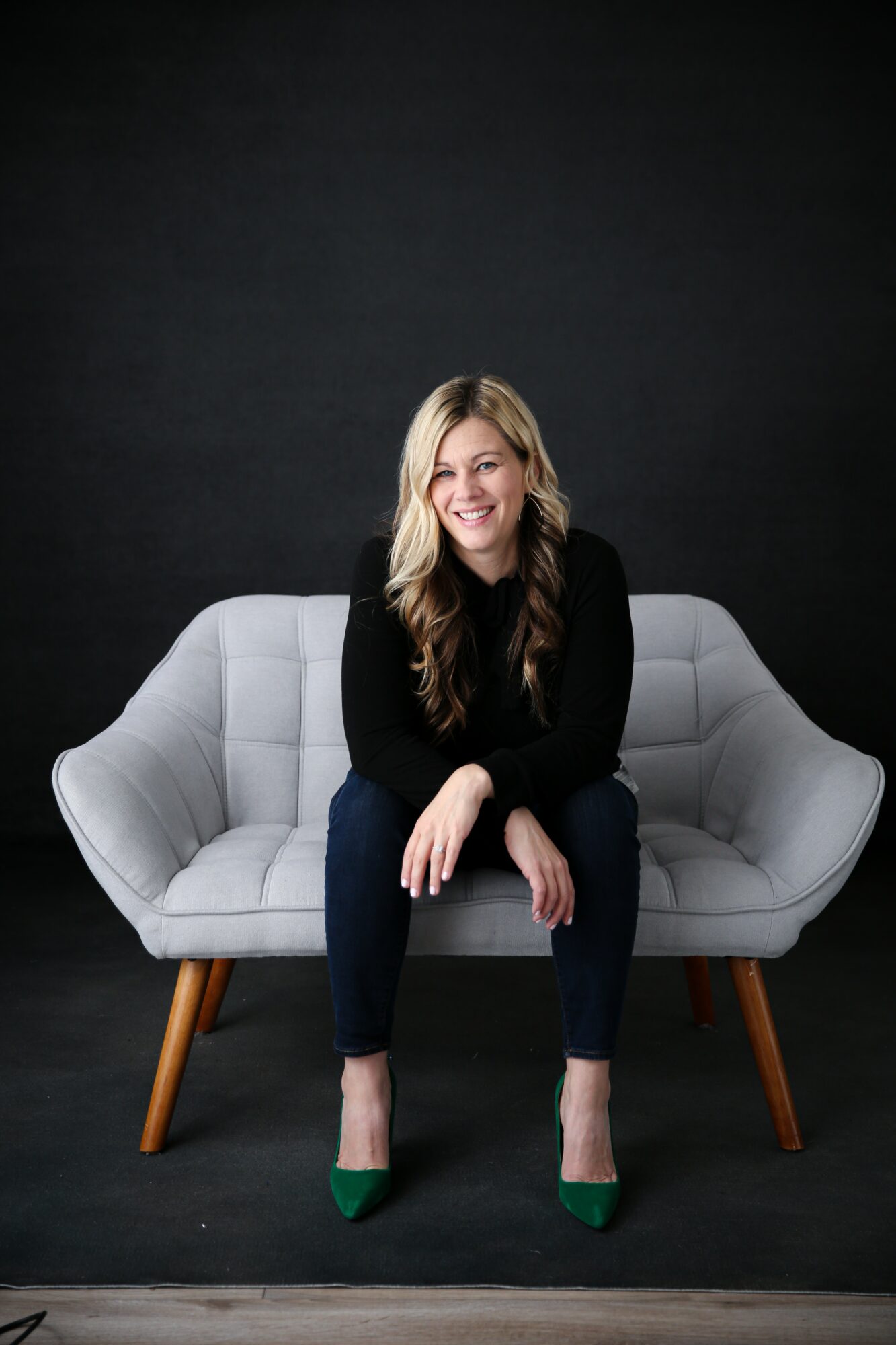 Woman with long blonde hair smiling, sitting on a light gray sofa against a dark background, wearing a black top, dark pants, and green shoes.