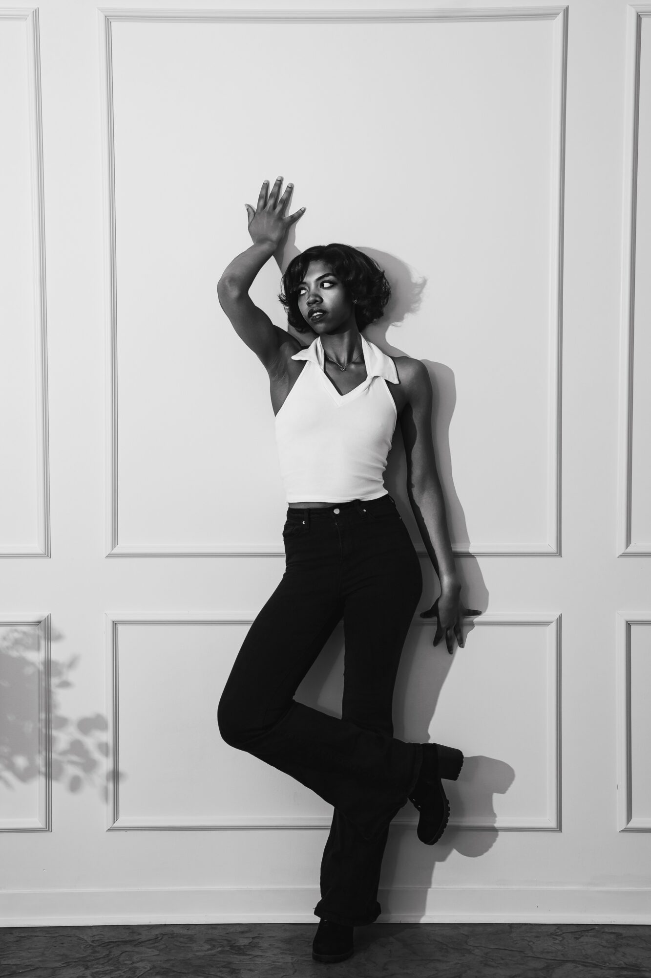 Woman standing against wall, raising her right hand, wearing a sleeveless top and dark pants, with her left leg crossed over her right.