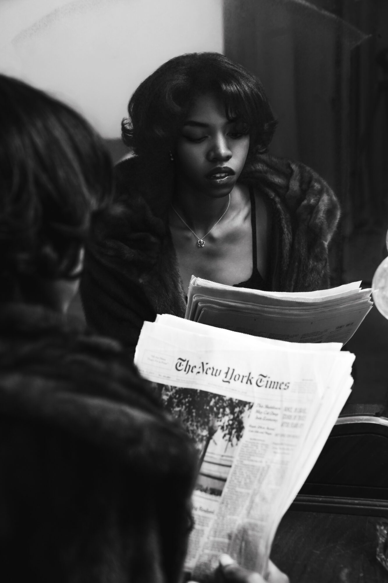 Two women reading newspapers, one with dark curly hair and the other with glasses, in a room with dark furniture.