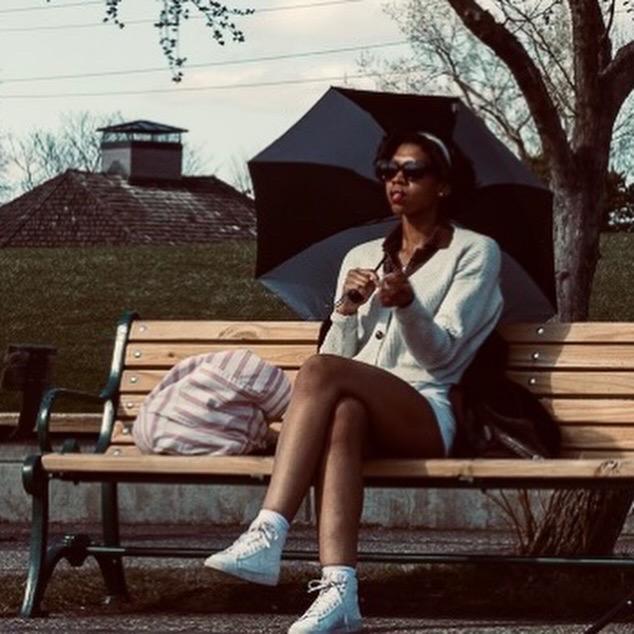 Woman sitting on a park bench holding a black umbrella, wearing sunglasses, white jacket, and sneakers, with a bag beside her.