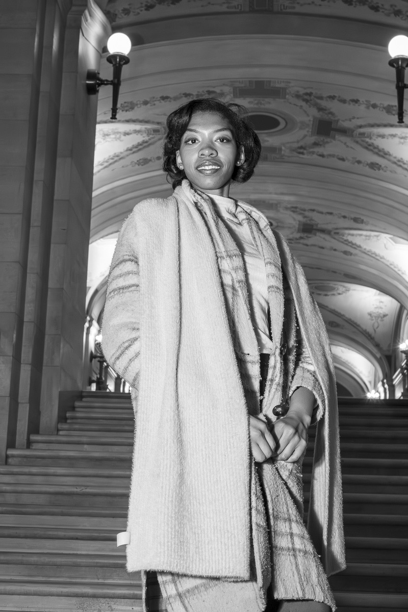 Woman standing indoors with ornate ceiling and stairs behind her, wearing a coat and scarf, smiling slightly.