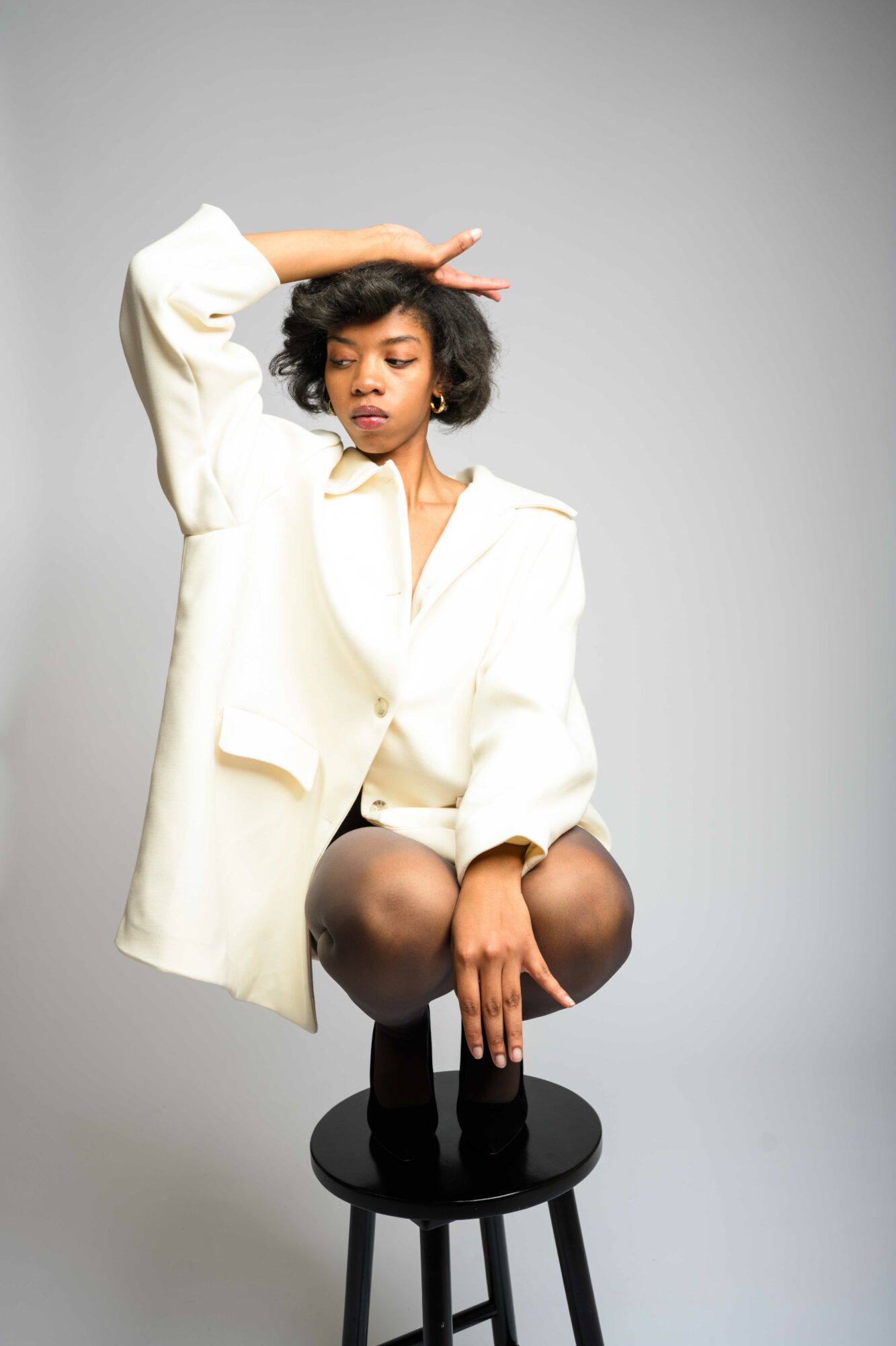 Young woman with curly hair sitting on a stool, wearing a white oversized blazer, with her hand on her forehead, against a plain gray background.