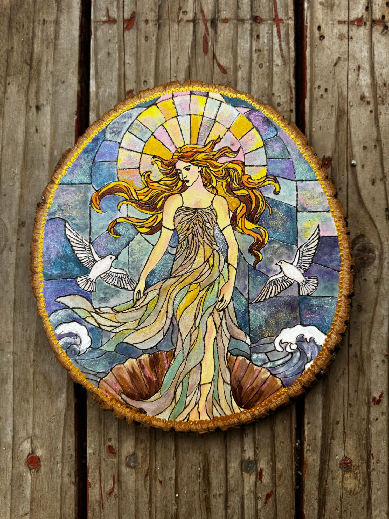 Colorful stained glass depiction of a woman with flowing hair, surrounded by birds and a scenic background, on a wooden surface.