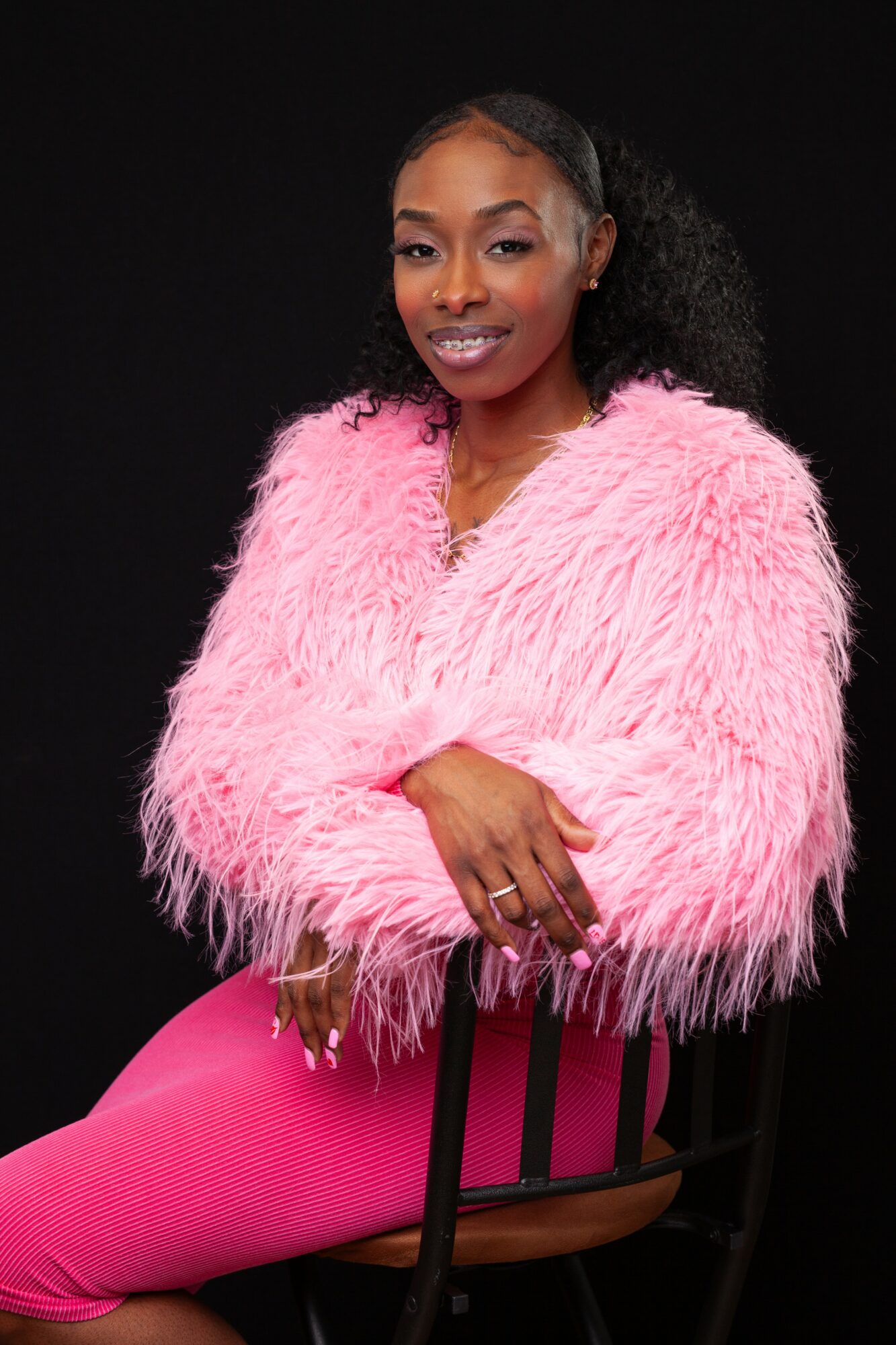Woman with curly hair in a pink fluffy jacket and matching pants, sitting on a chair against a black background.