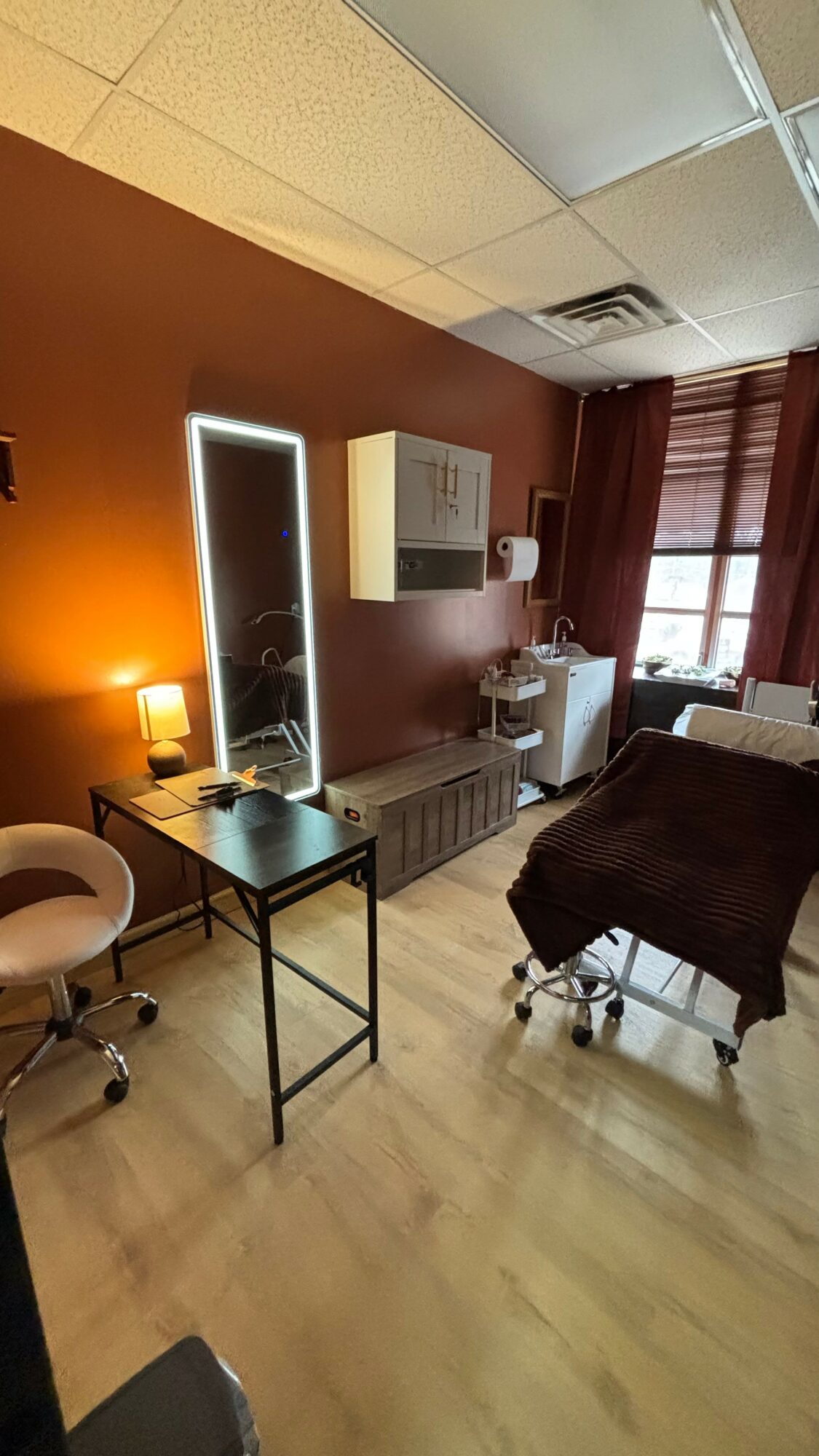 Room with a desk, mirror, cabinet, bed, and window with curtains, illuminated by warm lighting.