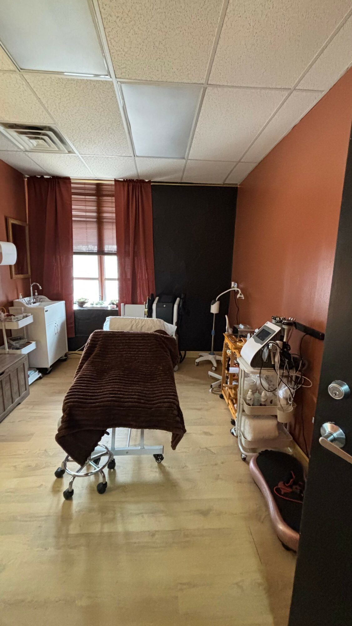 Treatment room with a bed, curtains, and medical equipment, walls painted in warm tones, and a window with natural light.