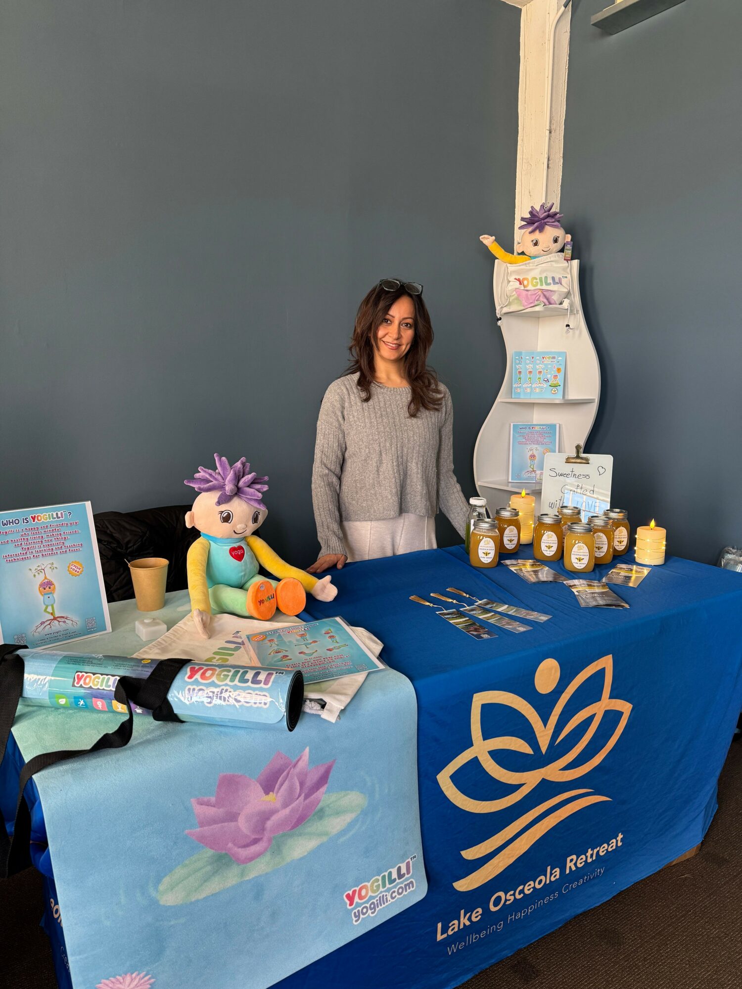 Woman standing behind a table with various items, including bottles and a purple flower, in a room with blue walls.