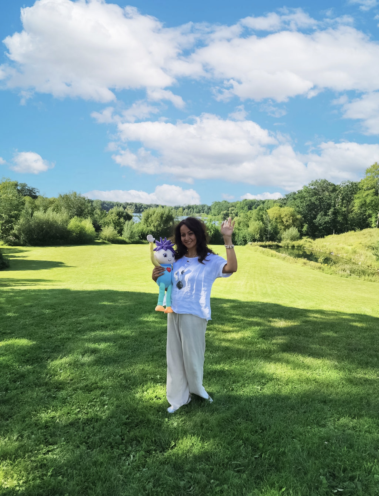Person standing on grass holding a puppet, with trees and blue sky with clouds in background.