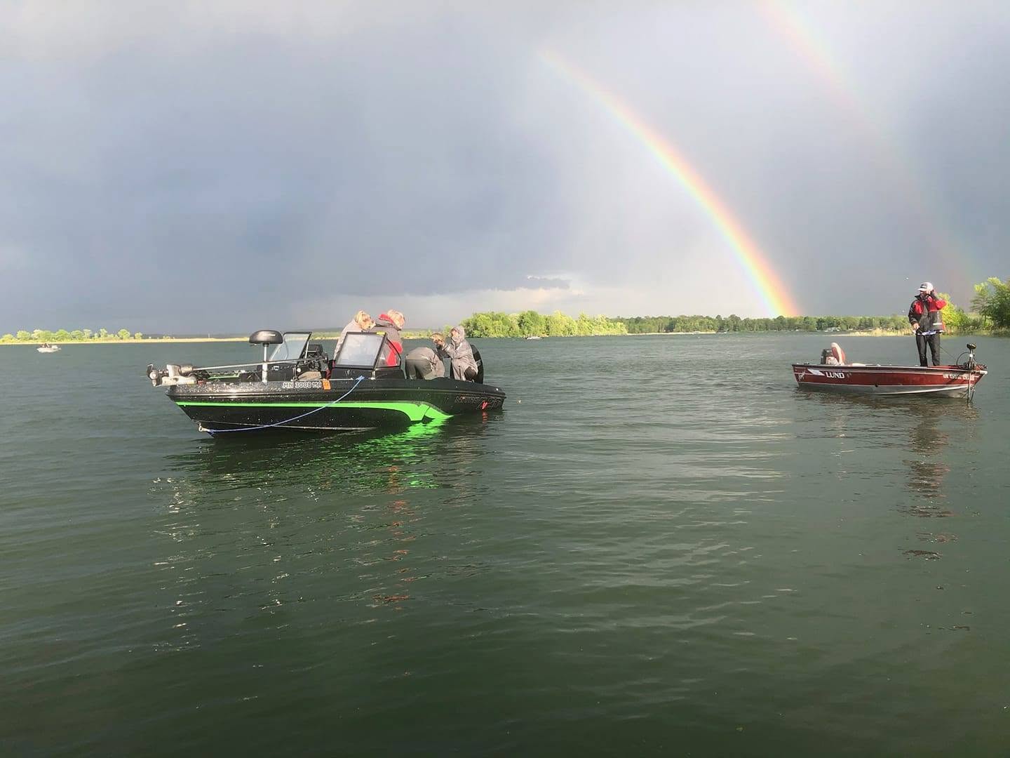 Two boats on water with a rainbow in cloudy sky, one person fishing on the right boat, calm water surface.