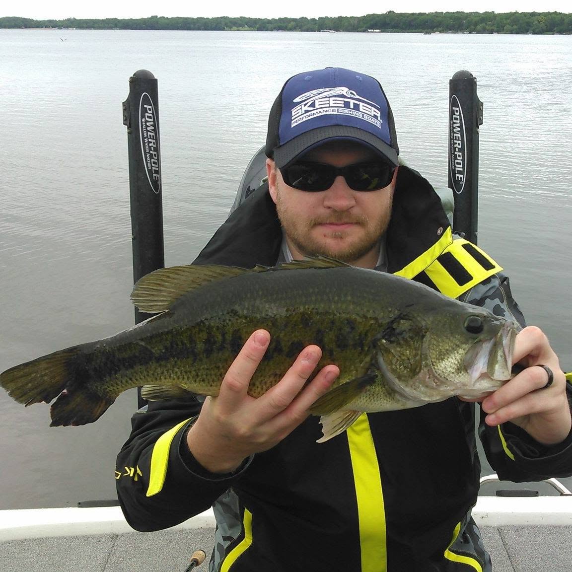 Man holding a large fish on a boat with water and distant shoreline in background.