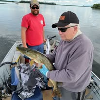 Two men on a boat, one holding a fish, the other standing nearby, water and trees in background.