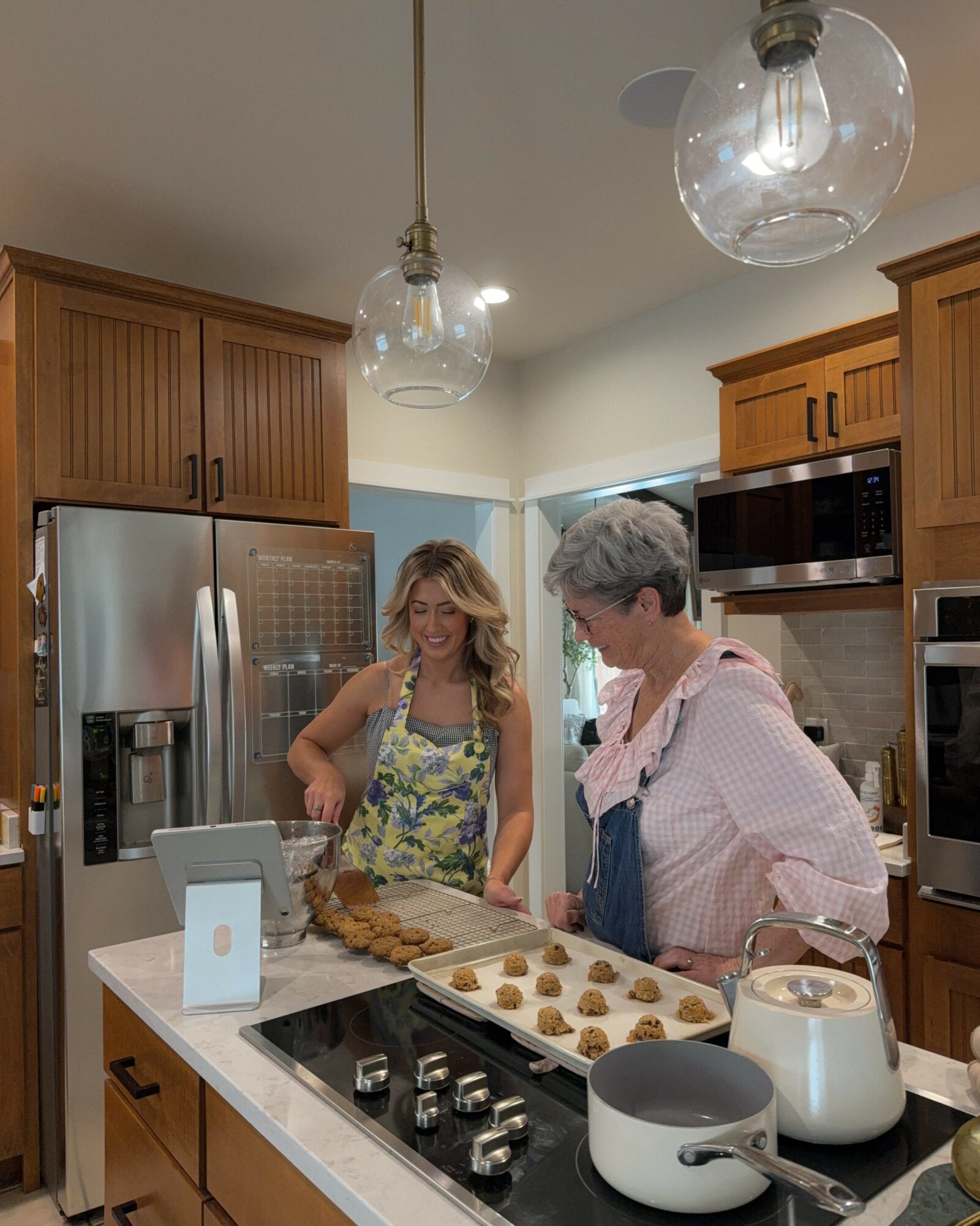 Two women baking cookies in a kitchen with wooden cabinets and modern appliances, smiling and talking, with baking tray on counter.