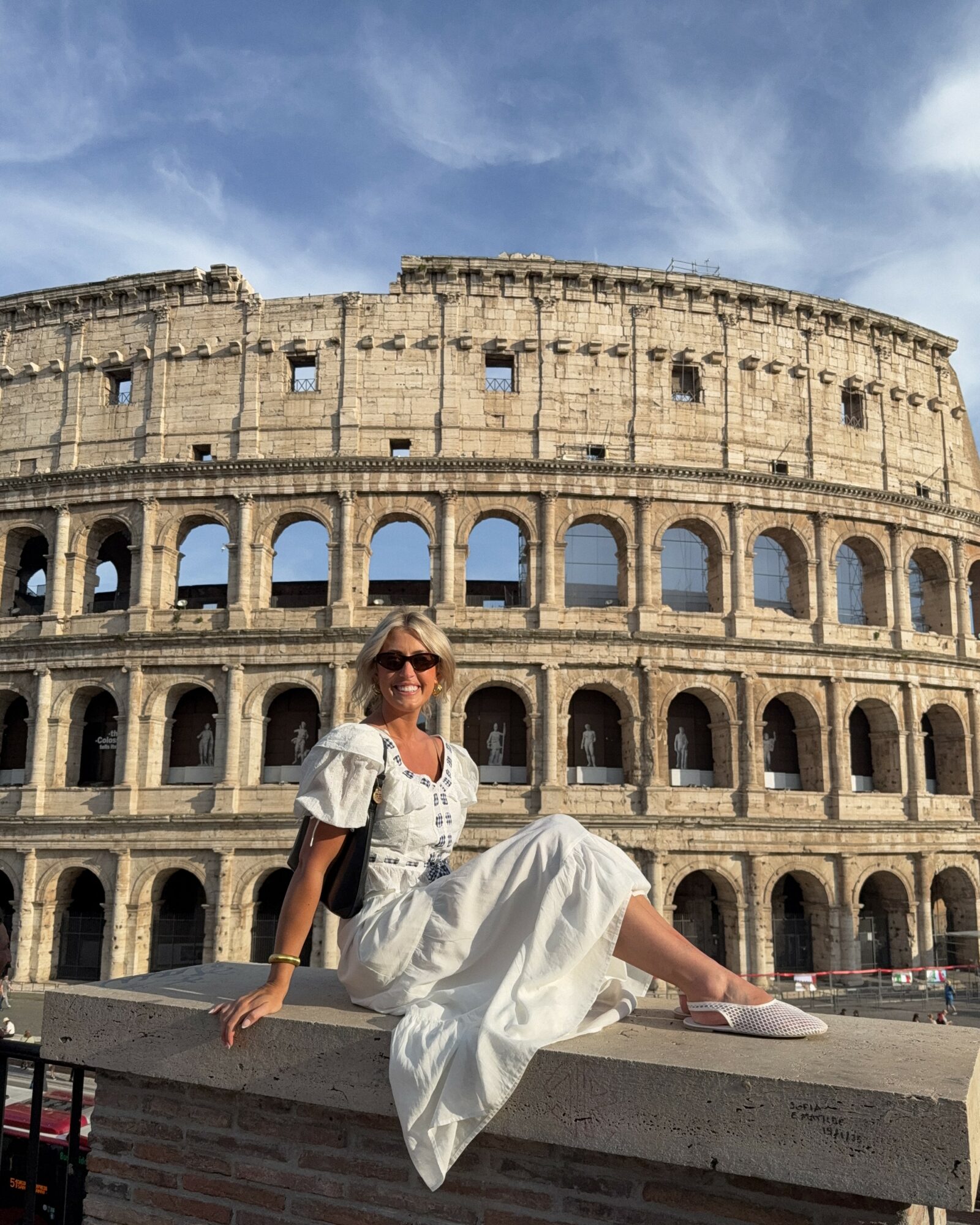 Woman sitting on a ledge in front of the Colosseum, wearing sunglasses and a white dress, smiling at the camera.