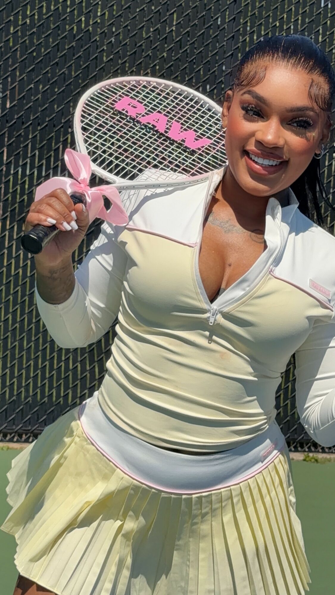 Woman holding a tennis racket, smiling, outdoors on a sunny day, wearing a light-colored jacket and pleated skirt.