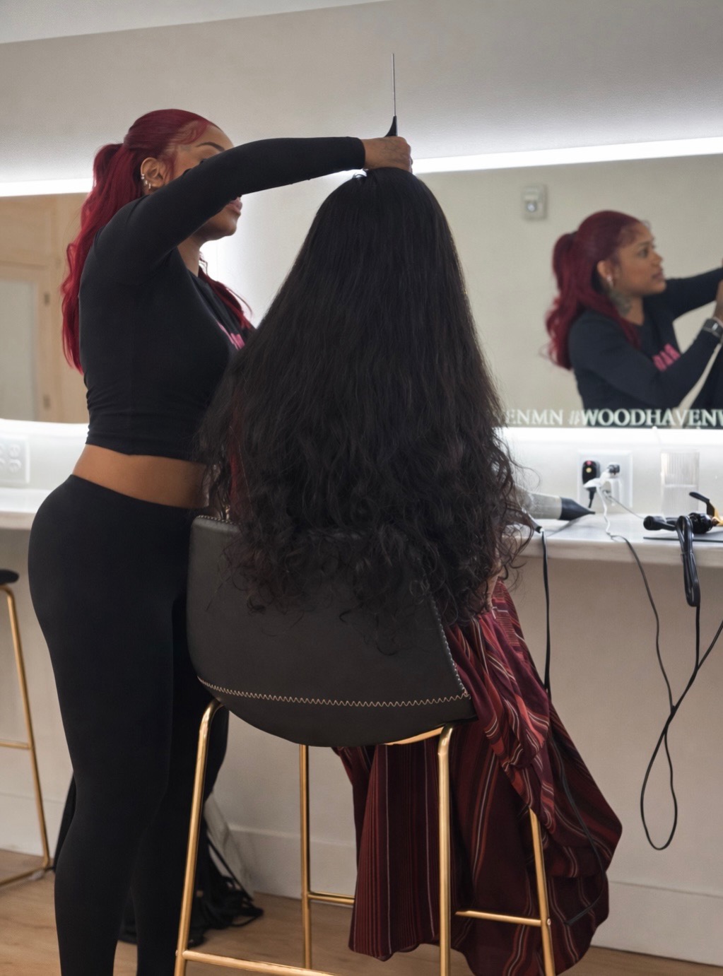 Person with long dark hair sitting in a salon chair, another person styling hair, mirror reflects the scene, modern salon setting.