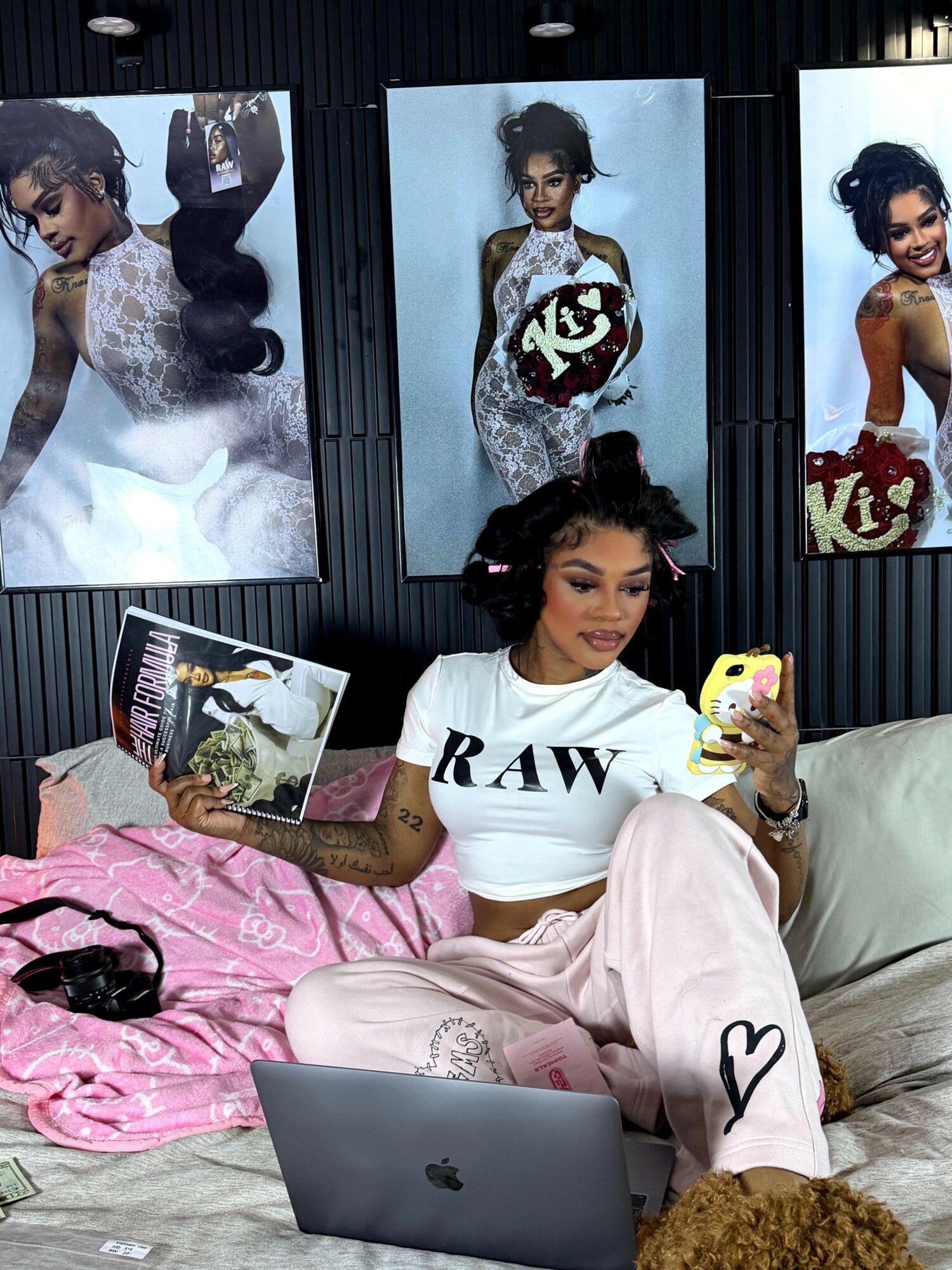 Young woman sitting on a couch, holding a magazine and a phone, with a laptop nearby and posters on the wall behind her.