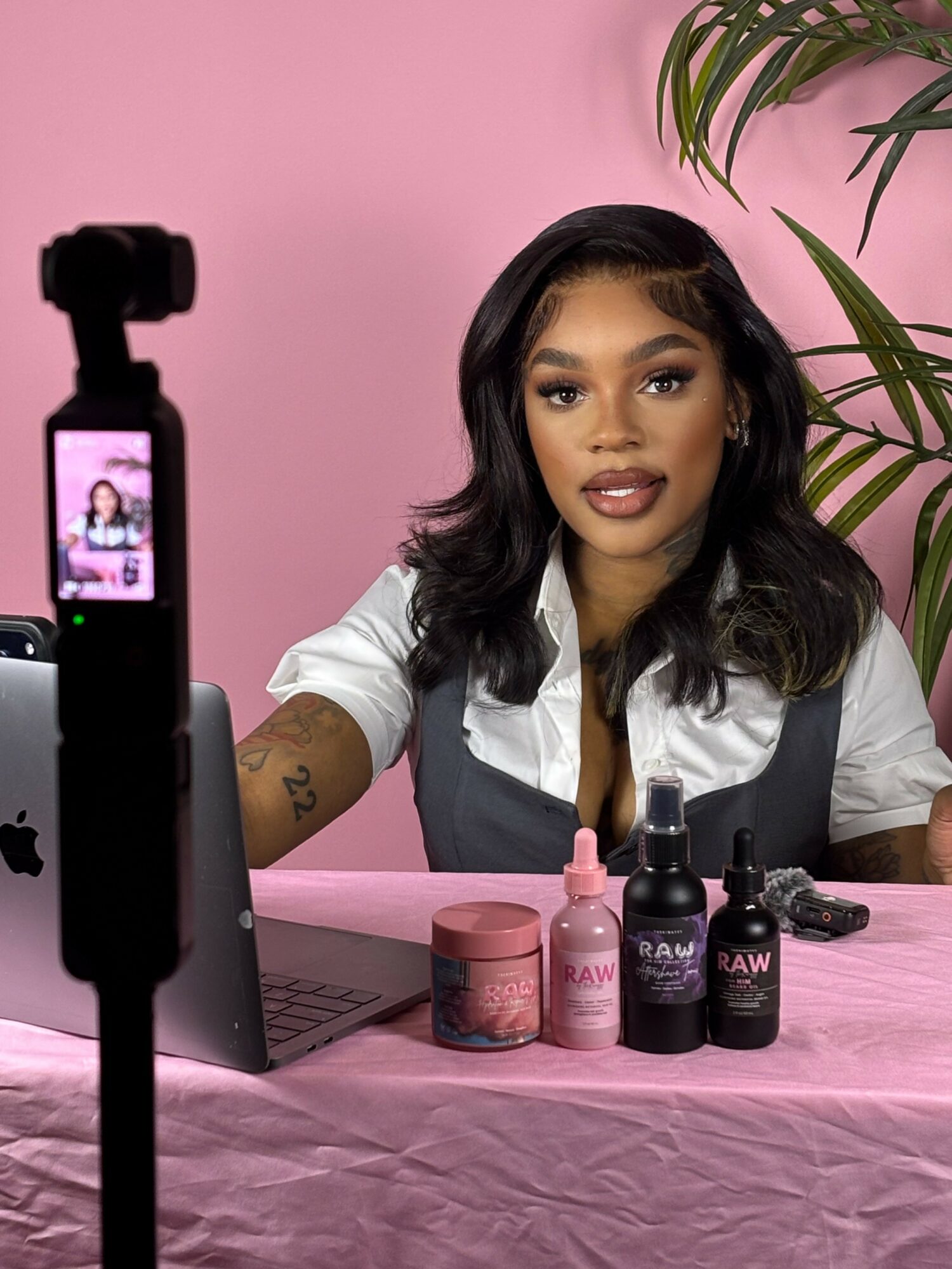 Woman with dark hair and makeup sitting at a table with skincare products, pink background, plant, and camera setup.