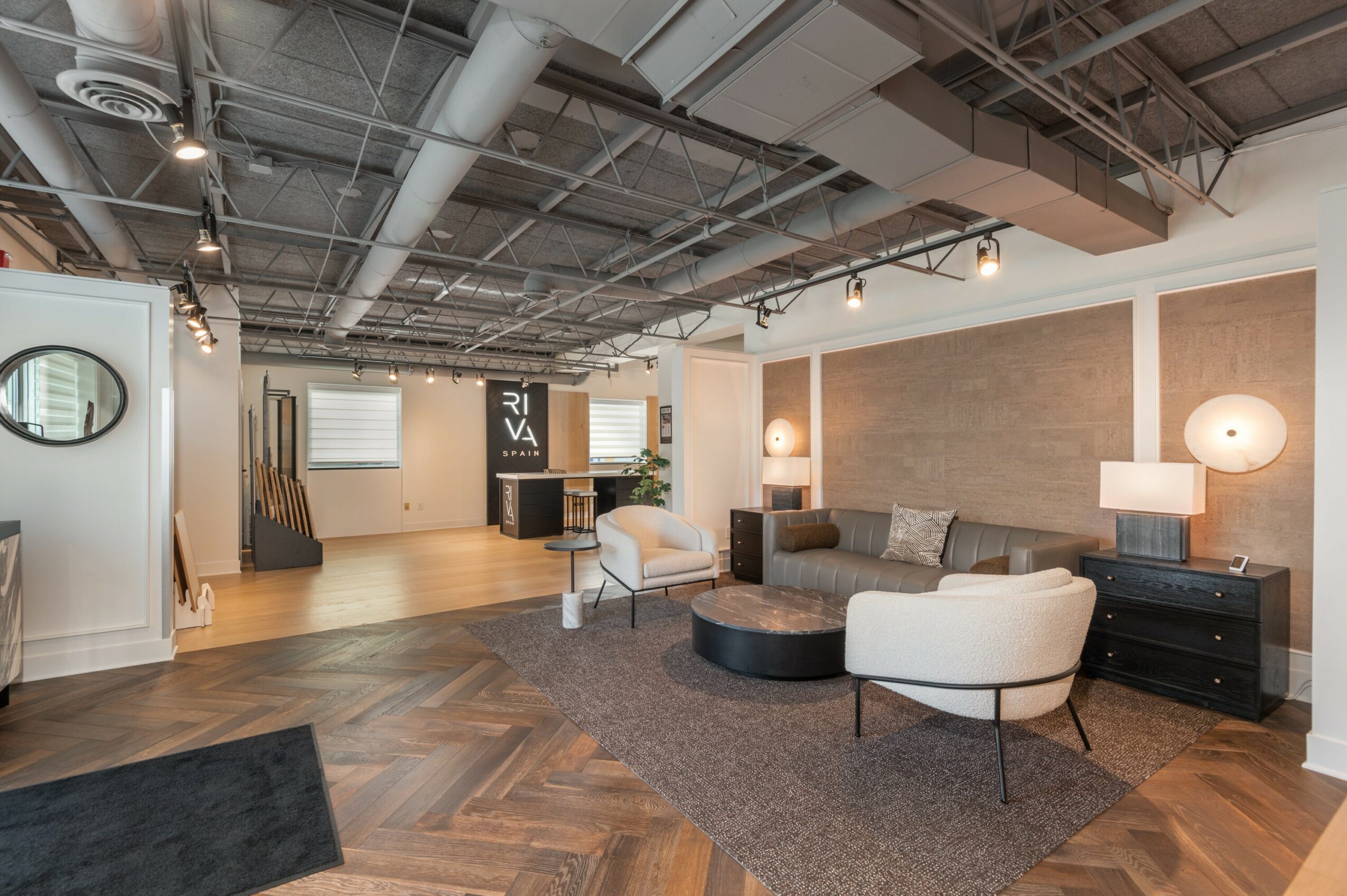 Modern living room with sofa, armchair, lamps, and wooden flooring, ceiling with exposed pipes, and a wall with a textured panel.