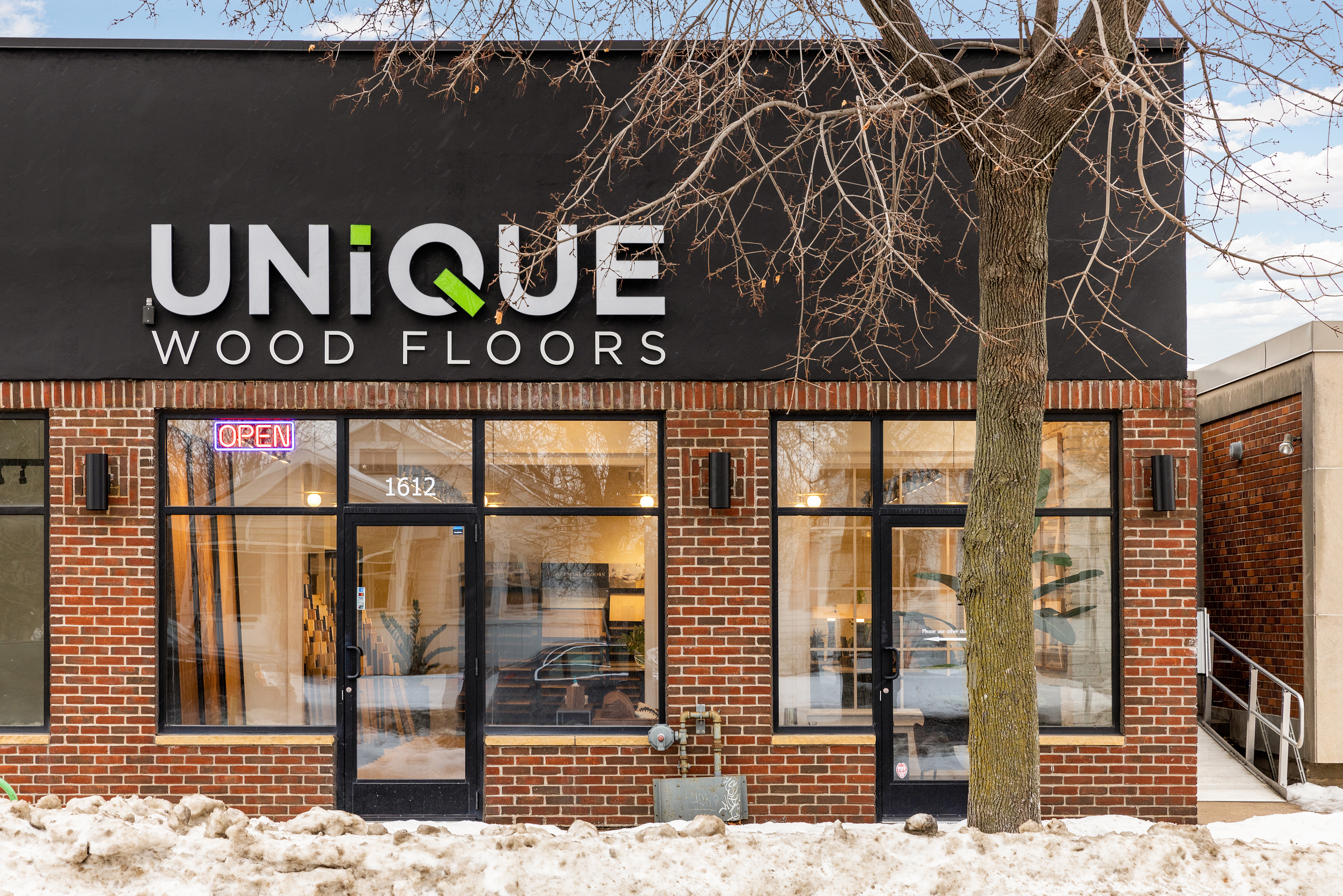 Storefront with large windows, brick exterior, and a sign reading 'UNIQUE WOOD FLOORS'. Tree in front, door with 'OPEN' sign.