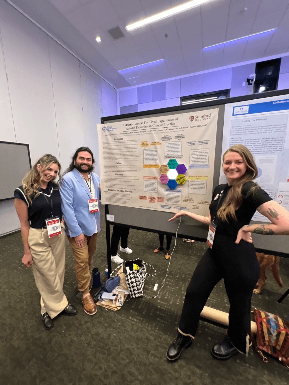 Three people stand in front of a research poster at a conference, smiling. The poster is on a black display board in a room with white walls and ceiling lights.