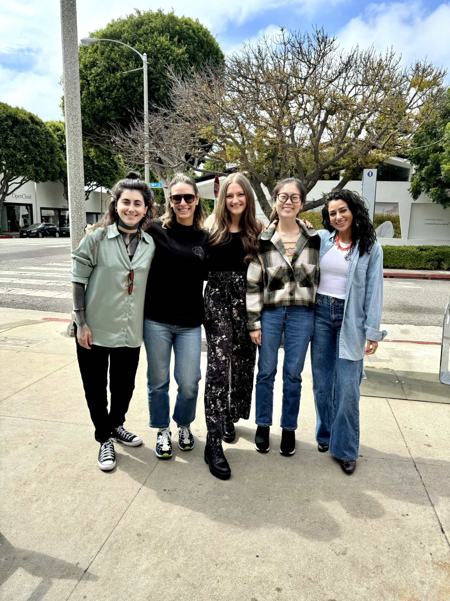 Five women standing together outdoors on a sidewalk, smiling, with trees and a street in the background.