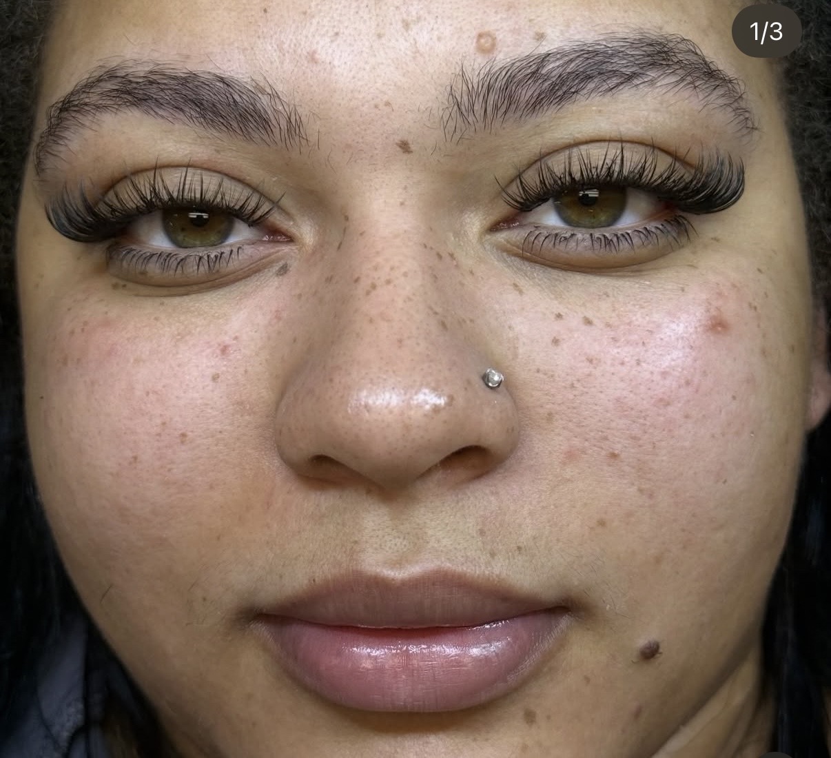 Close-up of a person's face with makeup, including long eyelashes, a nose piercing, and visible skin blemishes.