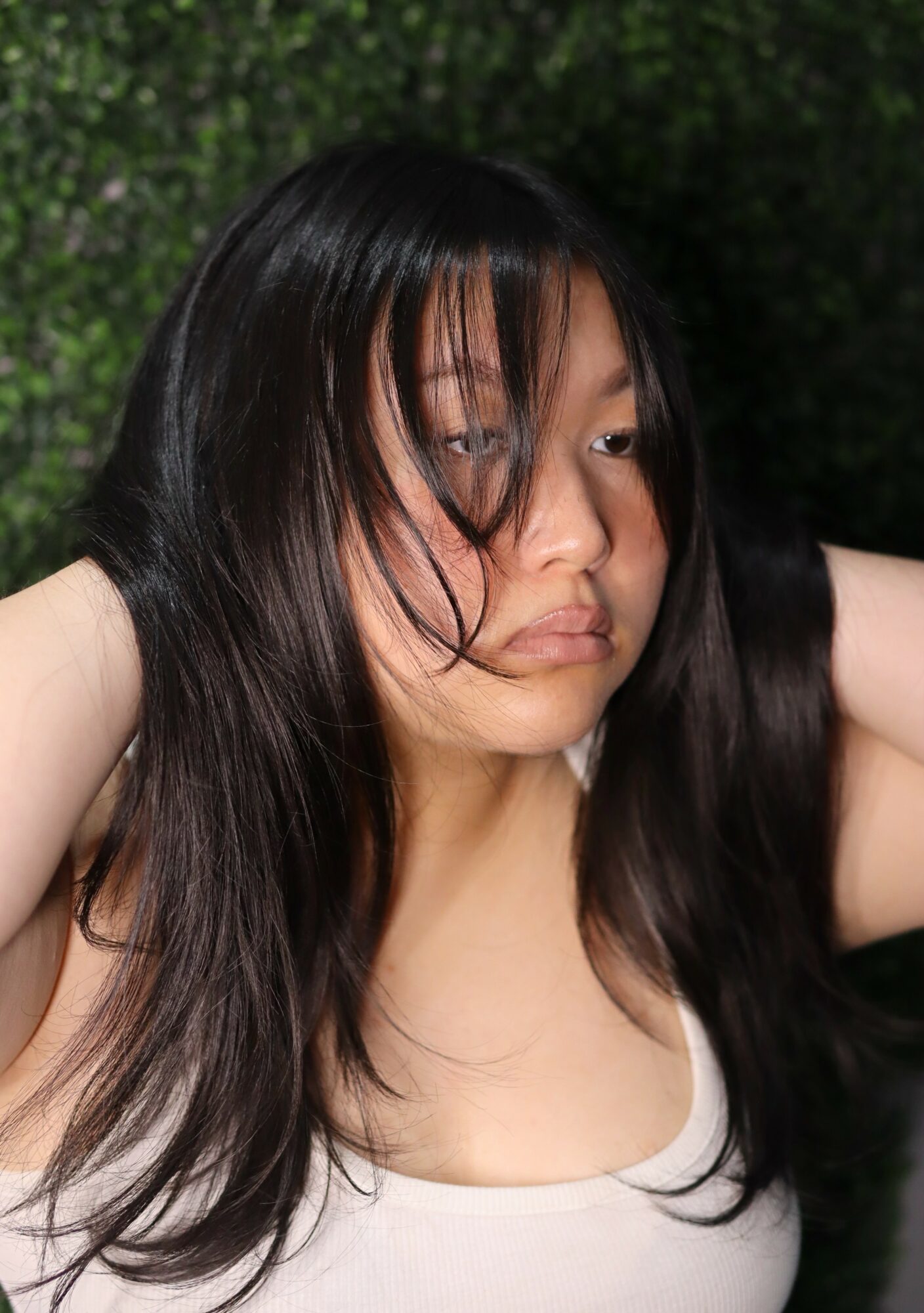 Young woman with long dark hair looking to the side outdoors, green background, wearing a white top.