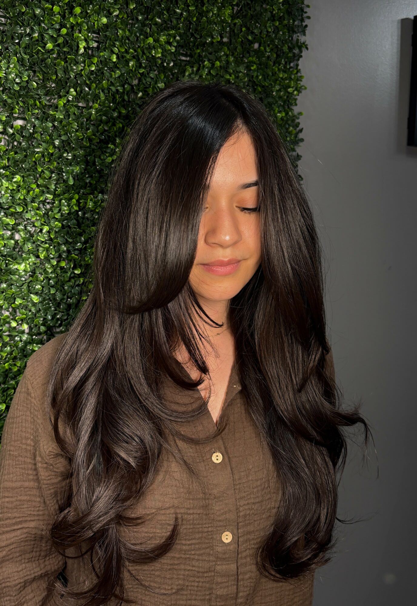 Young woman with long, wavy dark hair and closed eyes, standing in front of a green leafy background.