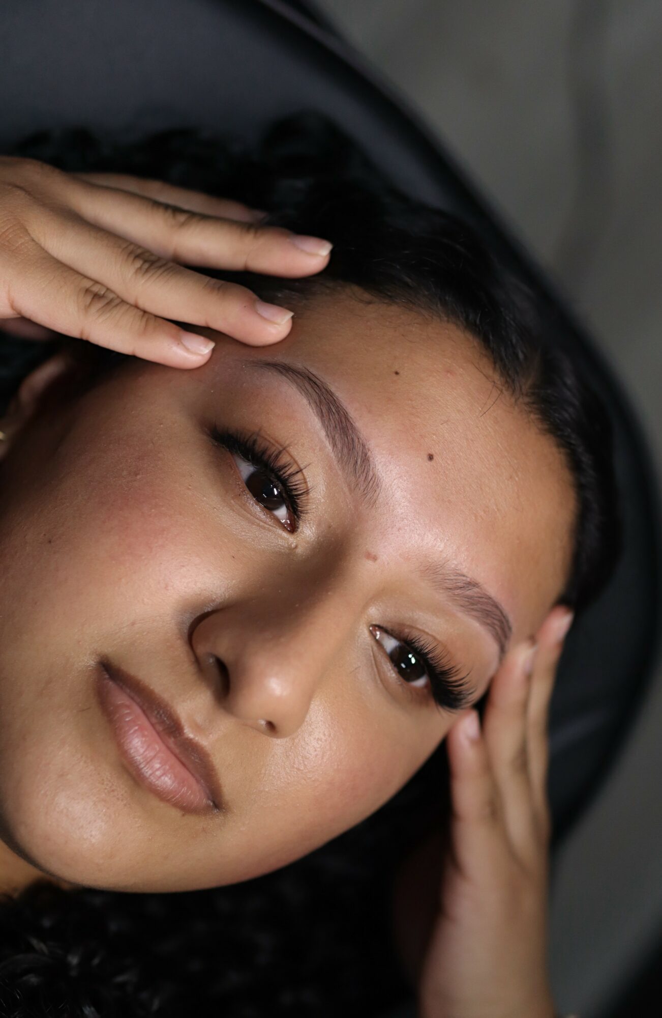 Close-up of a woman with hands near her face, looking at the camera, with dark hair and makeup.