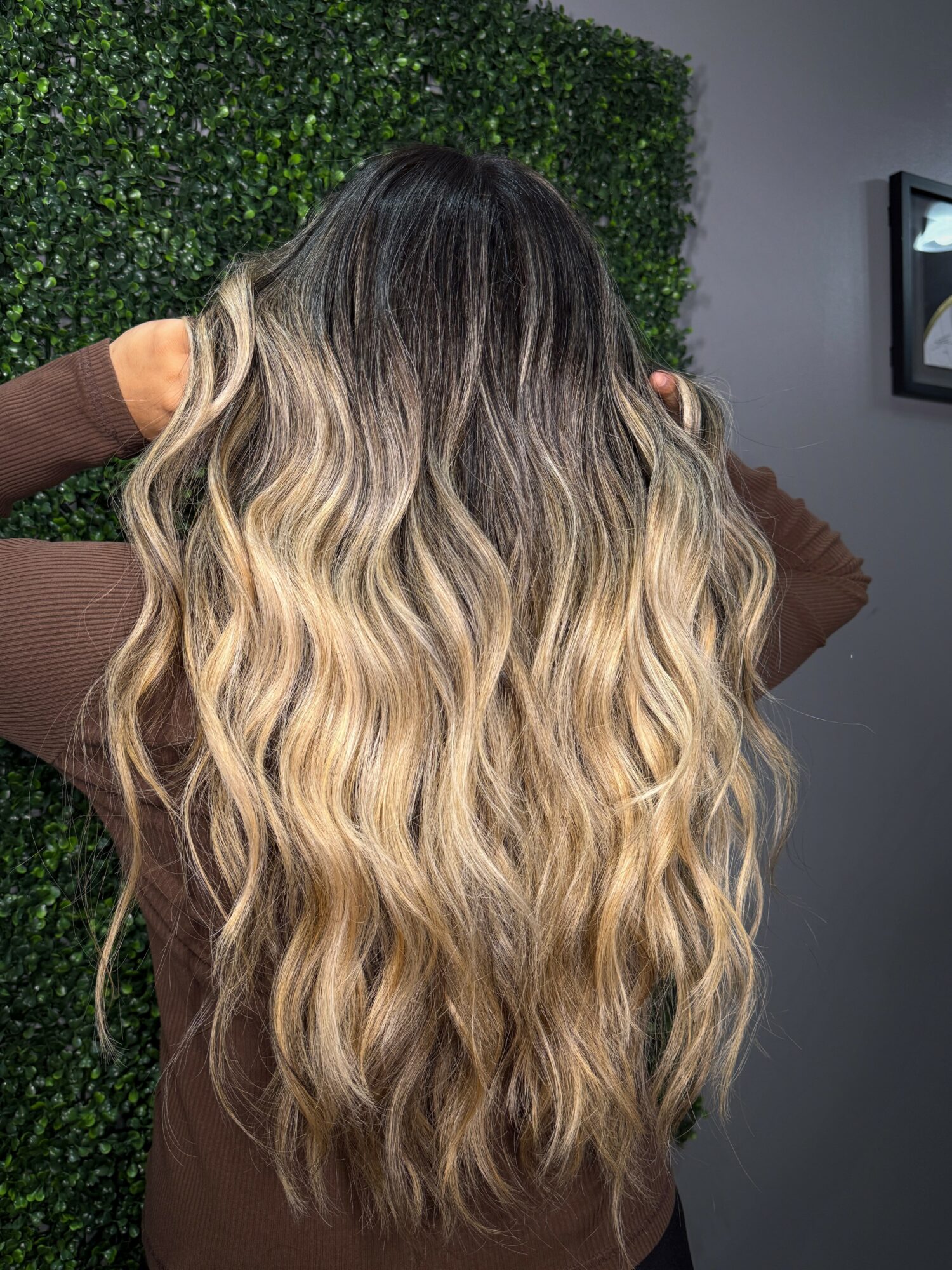 Back view of a woman with long, wavy hair, standing indoors near a green wall and a gray wall.