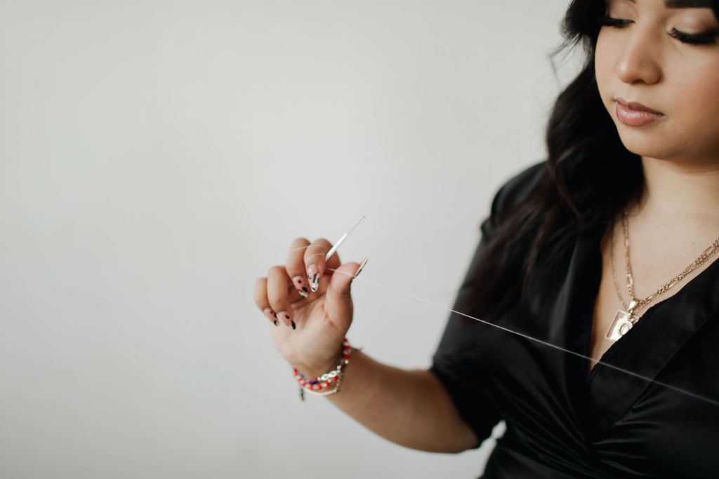 Young woman with dark hair and jewelry, wearing a black top, holding a string or thread, against a plain background.