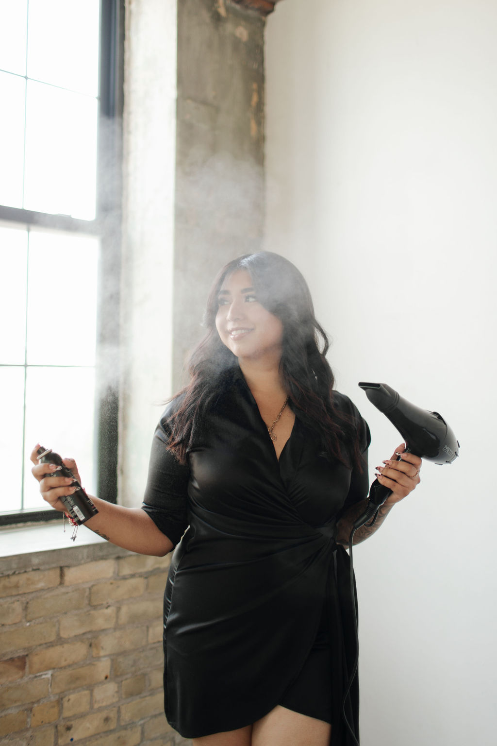 Woman in black dress holding a spray bottle and cloth, smiling near a window with sunlight streaming in.