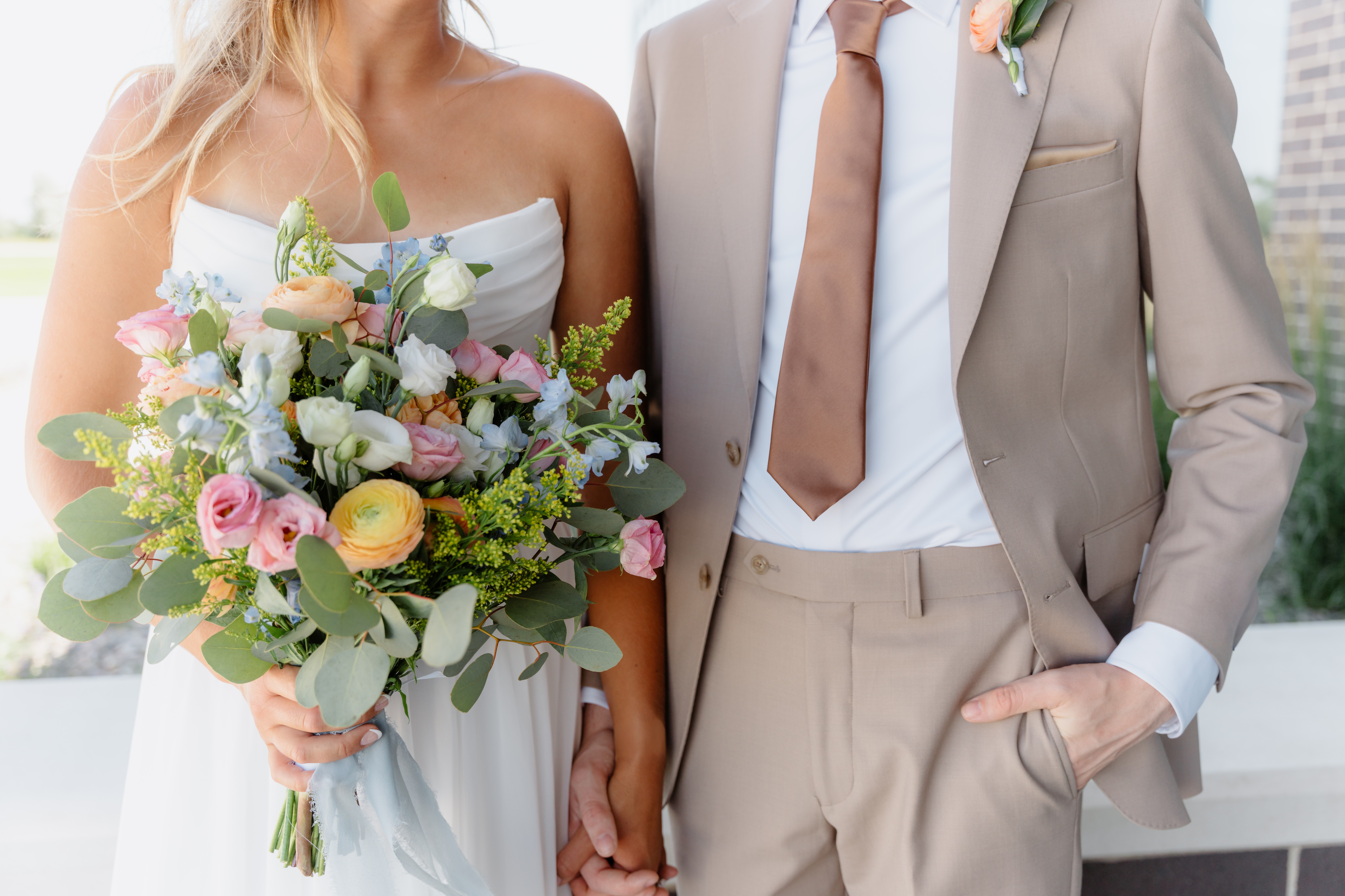 Bride holding a bouquet of flowers, wearing a white dress, with a groom in a beige suit holding her hand.