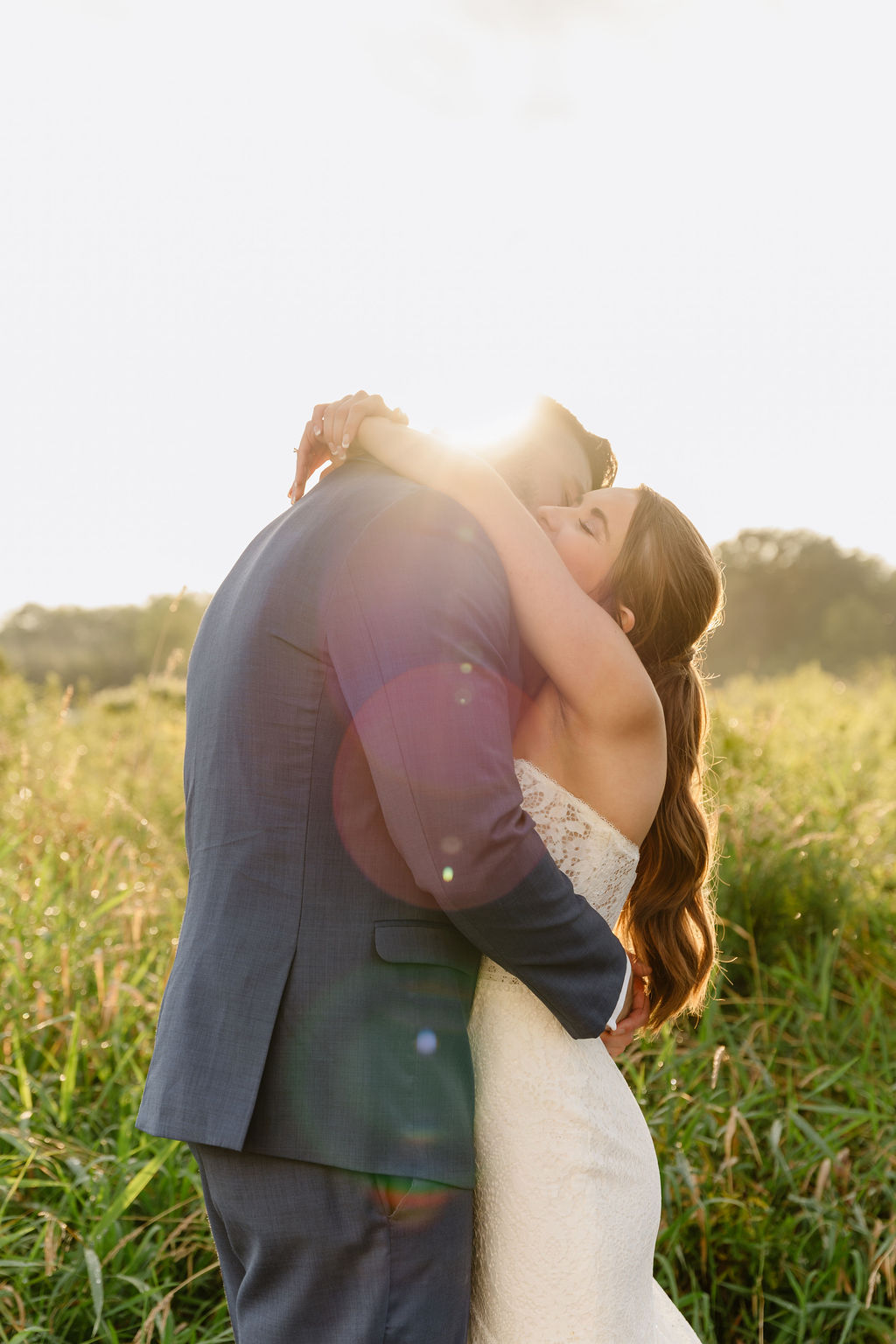 A couple embraces outdoors in a grassy field with sunlight behind them, the woman in a white dress and the man in a suit.