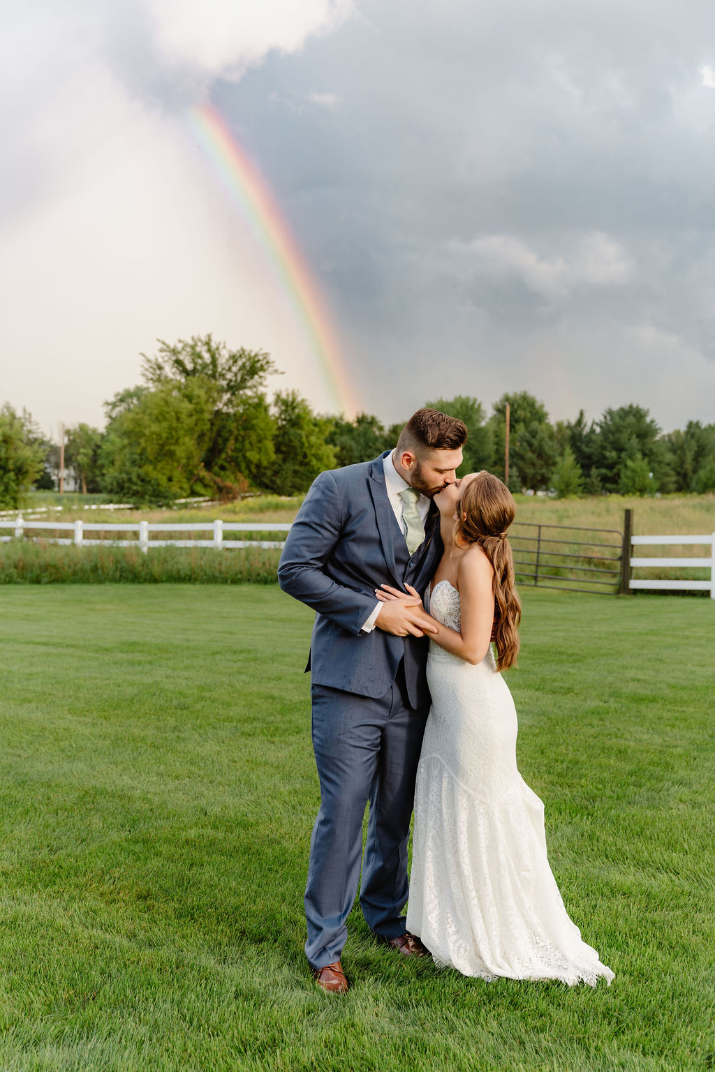 A couple in wedding attire standing on a grassy field, with a rainbow and cloudy sky in the background.