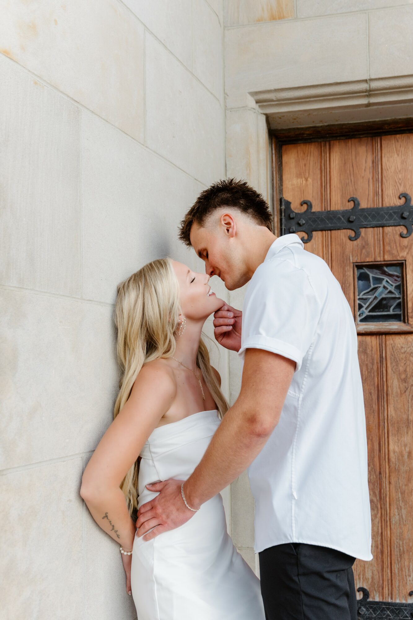 A couple stands close to each other, smiling and leaning in for a kiss against a stone wall and wooden door.