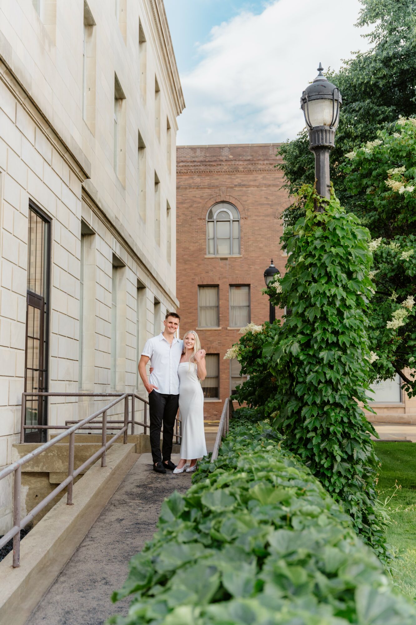 Two people stand on a sidewalk next to a building, with a lamppost and greenery nearby, under a partly cloudy sky.