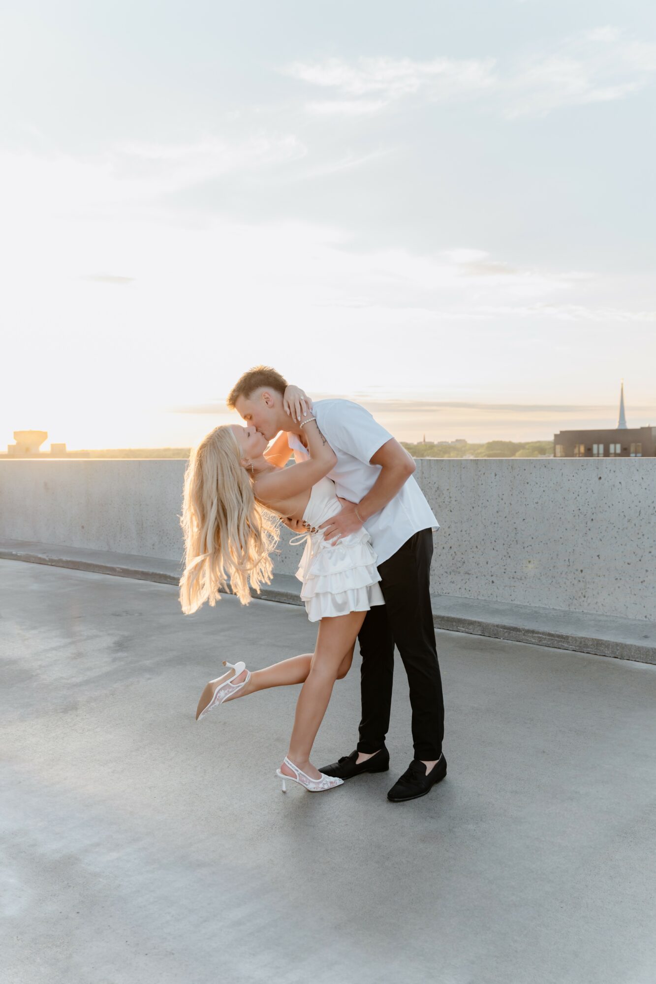 A couple is kissing on a rooftop with a cityscape and sky in the background, the woman is lifted off the ground.