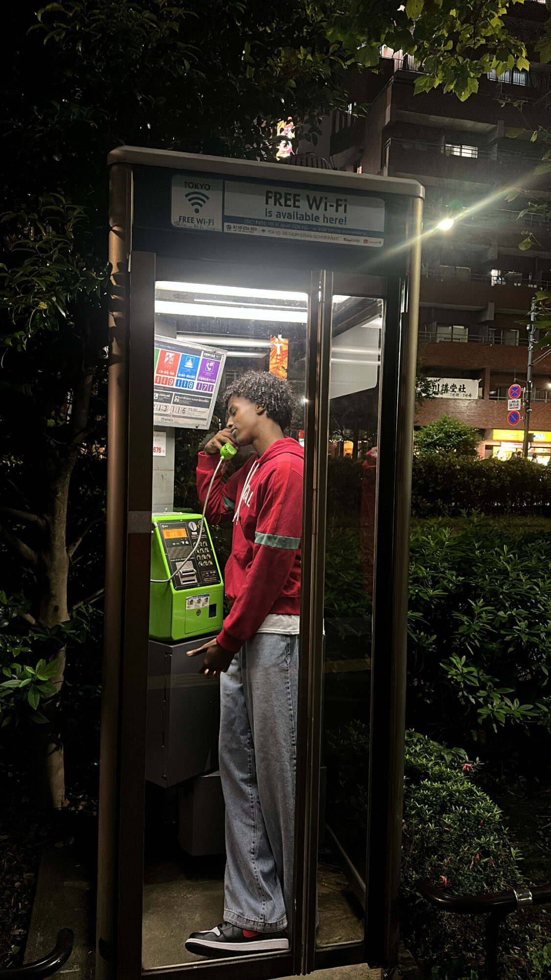 Person using a green ATM inside a phone booth at night, with trees and buildings in the background.