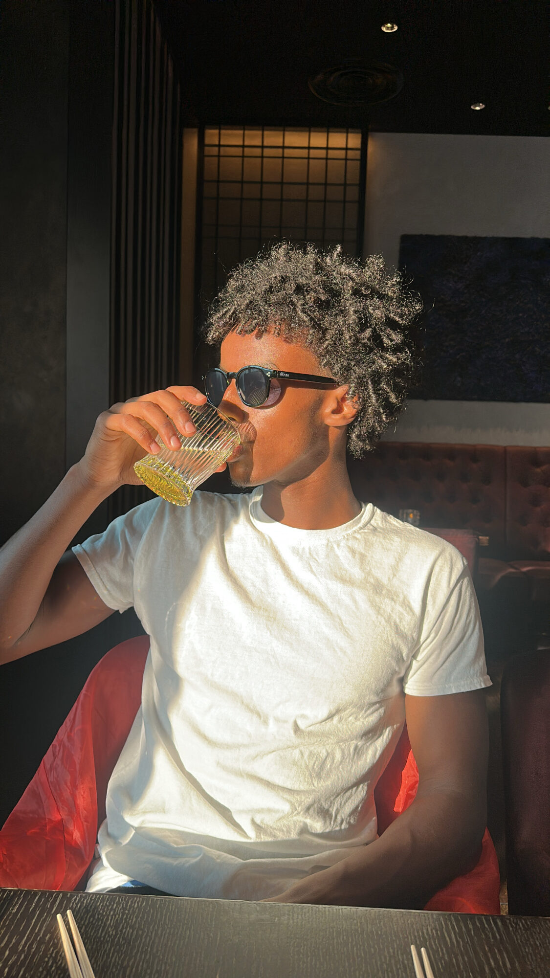 Person with curly hair wearing sunglasses and a white t-shirt, drinking from a glass, sitting indoors.