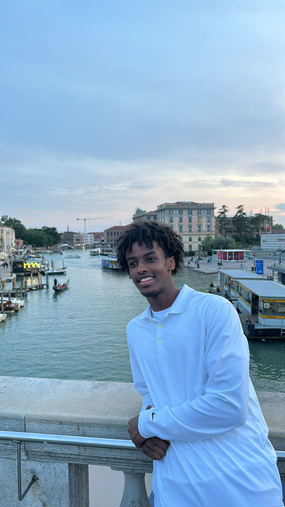 Young person with curly hair smiling, standing by a river with boats and buildings in the background.