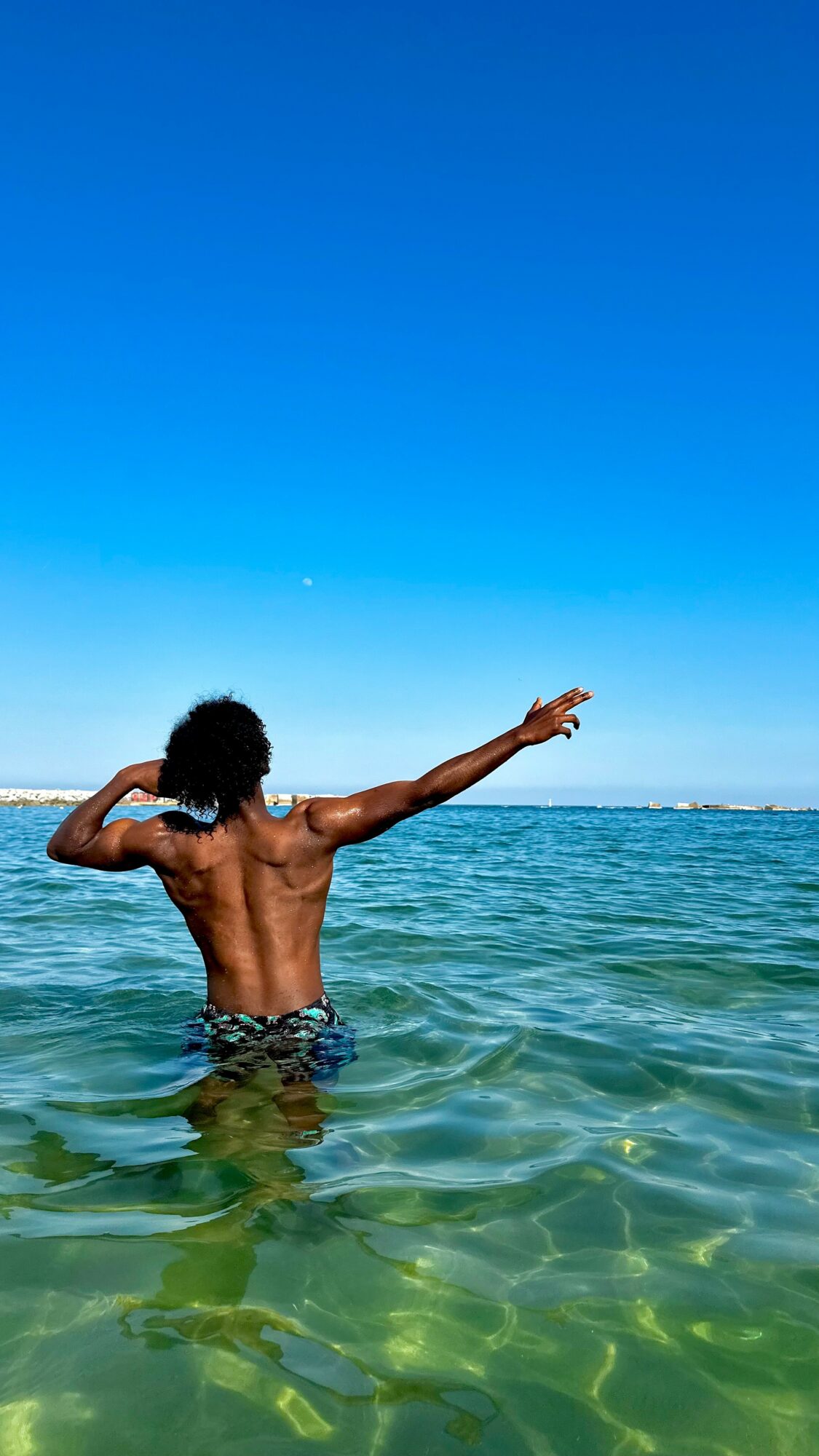Person with curly hair swimming in the ocean, arm extended, under a clear blue sky.