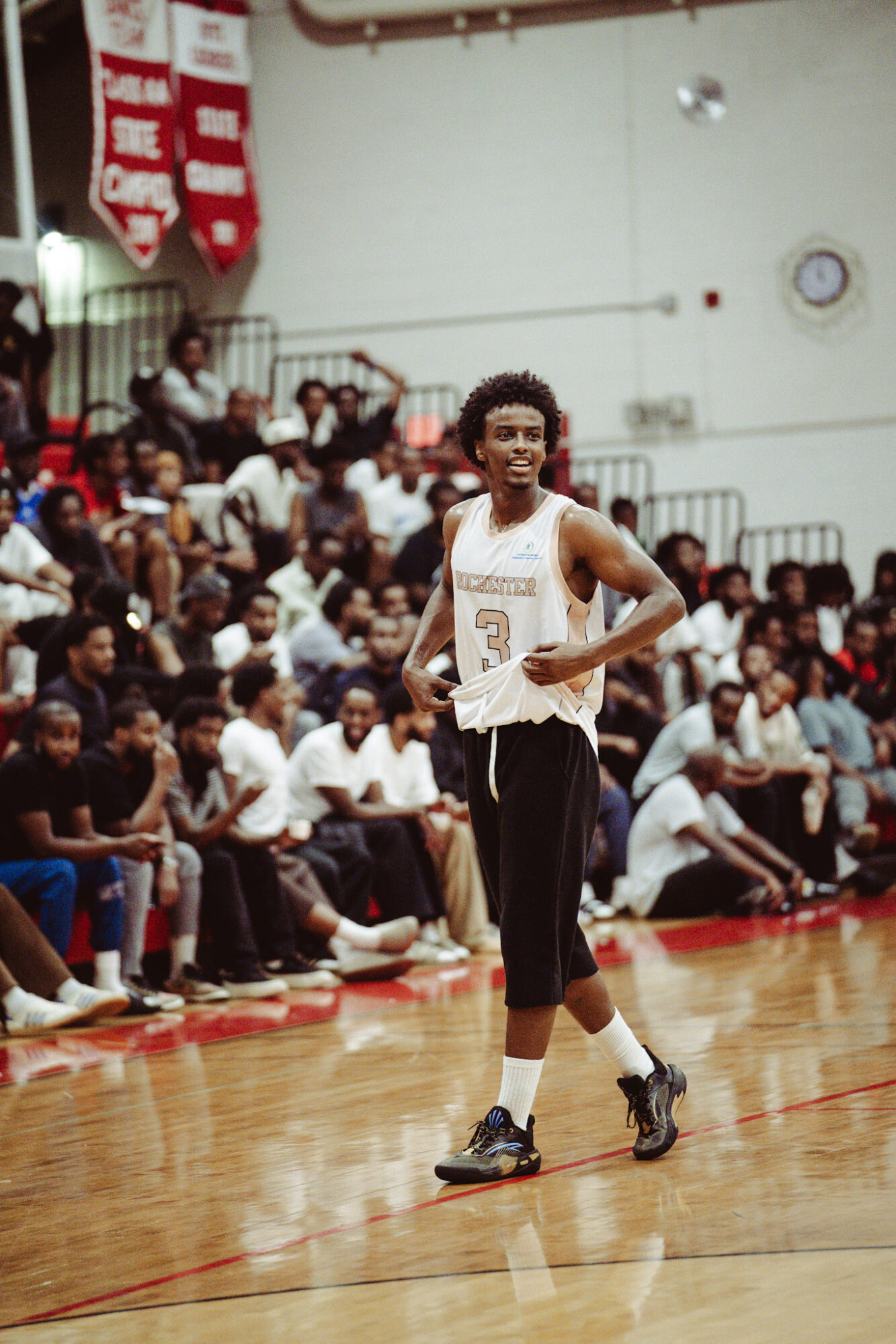 Basketball player with curly hair stands on court, crowd seated behind, gymnasium setting, player in white jersey and black pants.