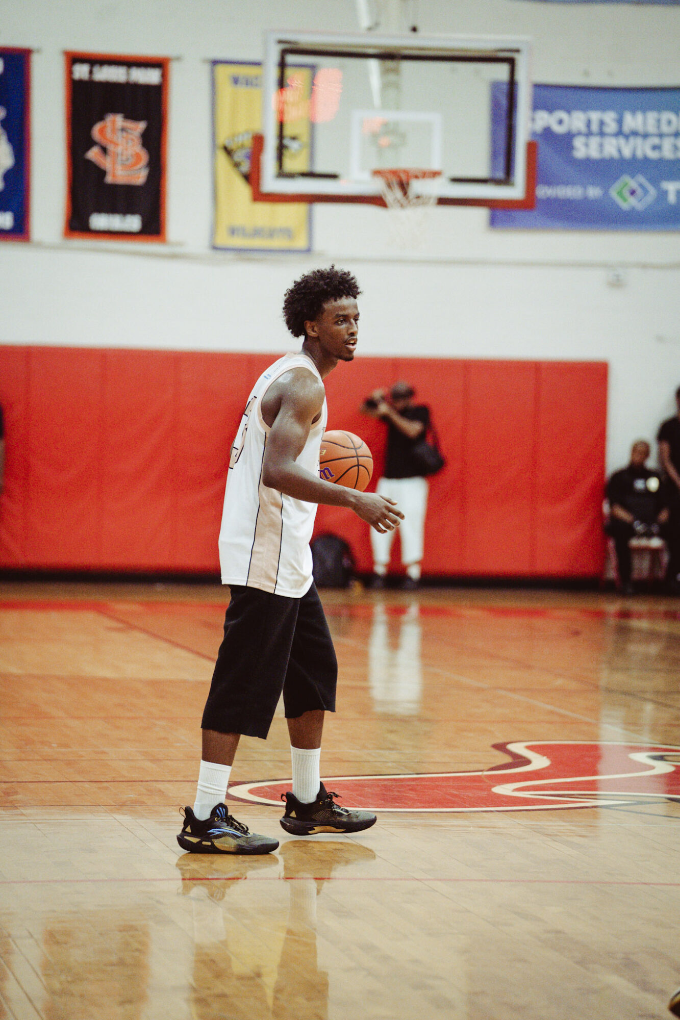 Young man in white tank top and black shorts holding a basketball on a gym court.
