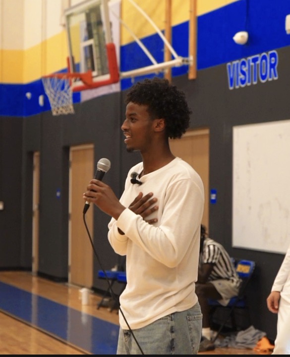 Young person speaking into a microphone in a gymnasium with a basketball hoop and a whiteboard behind.