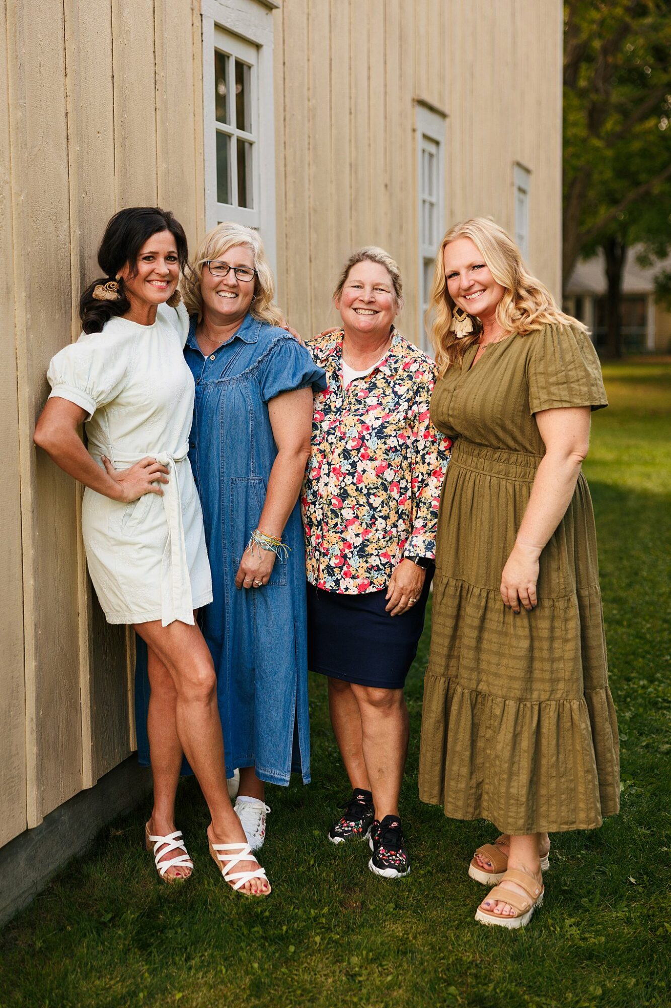 Four women stand outdoors near a beige building, smiling and posing for a photo, with trees in the background.