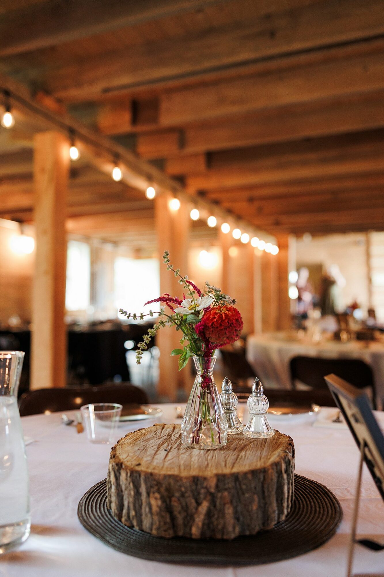 Decorative flower arrangement in a glass vase on a wooden slab at a rustic indoor event, string lights overhead.