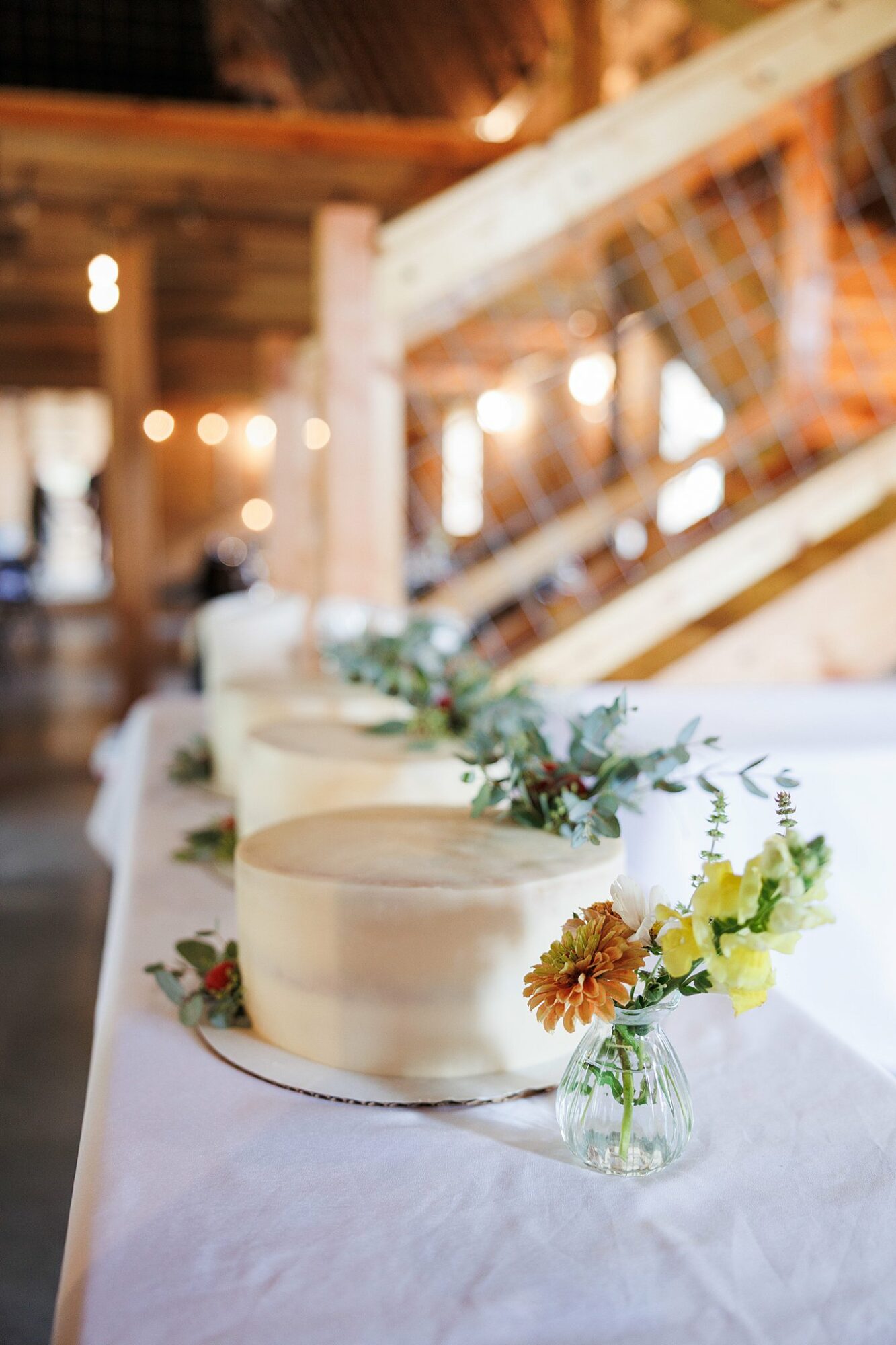 Three white cakes on a table with floral decorations, in a rustic indoor setting with wooden beams and warm lighting.